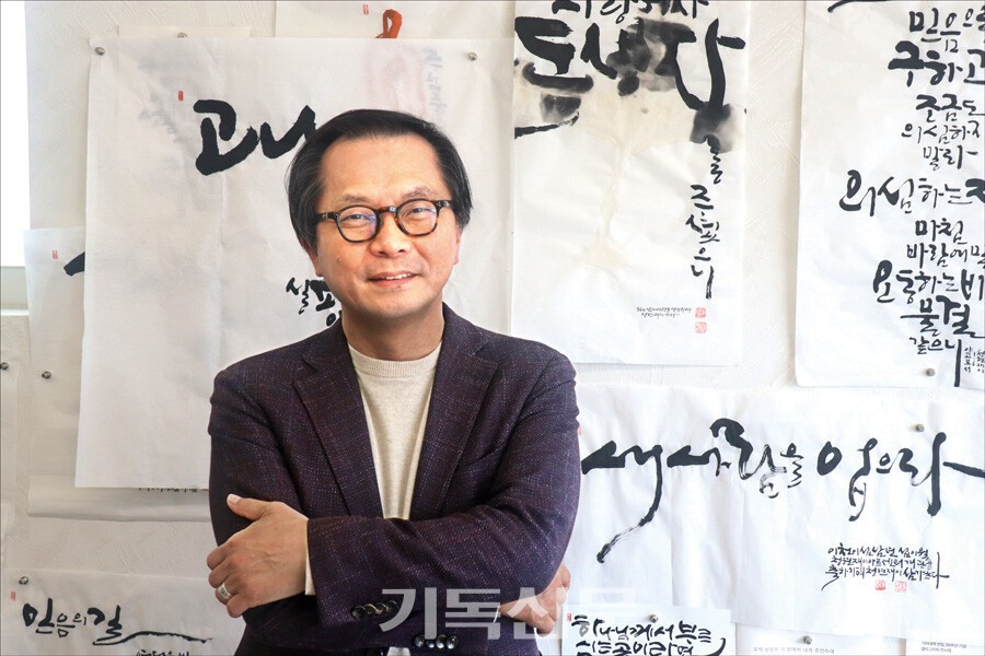 임동규 대표가 말씀 깃발전의 중요성을 설명하며, 벽 한쪽에 걸린 묵상의 결과물이 적힌 말씀을 소개하고 있다. 임 대표는 말씀깃발이 복음의 언어가 되는 매개체라고 강조한다.